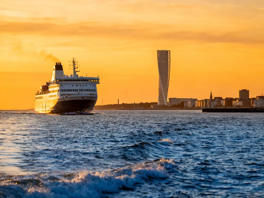 Nowoczesny prom pasażerski zbliżający się do portu w Malmö o zachodzie słońca na tle panoramy miasta i wieżowca Turning Torso.